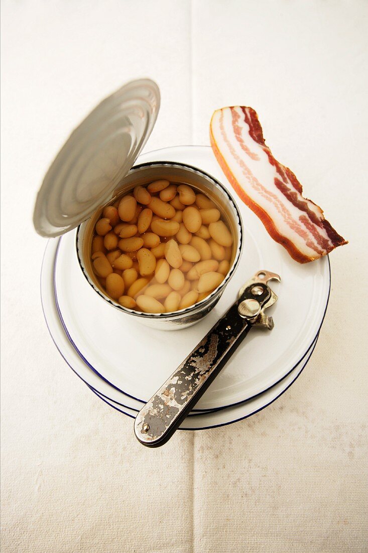 A tin of baked beans on a plate with bacon and a tin opener