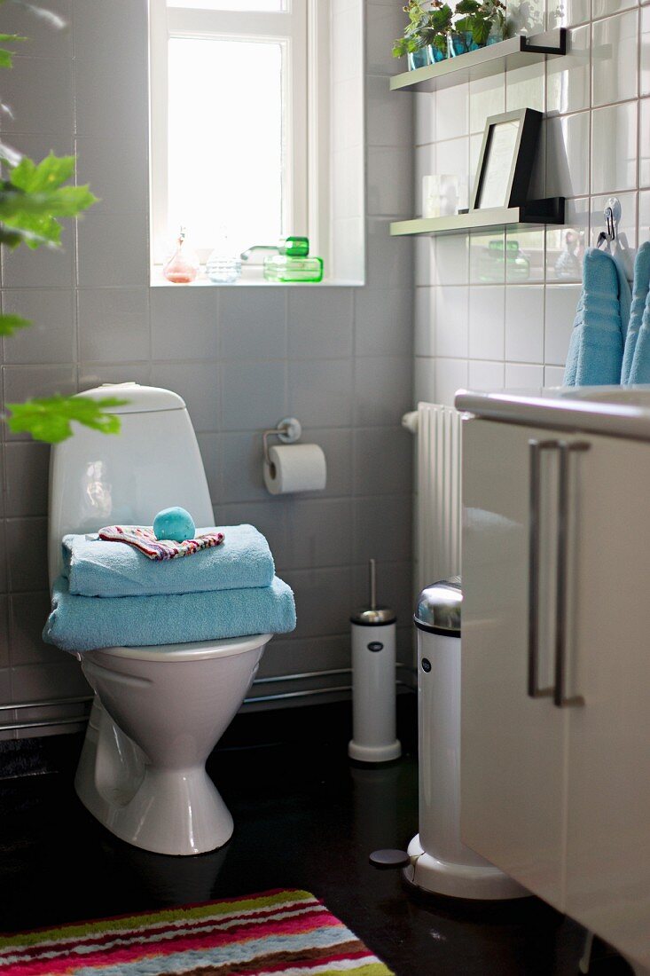 White-tiled bathroom with stack of towels on toilet below window