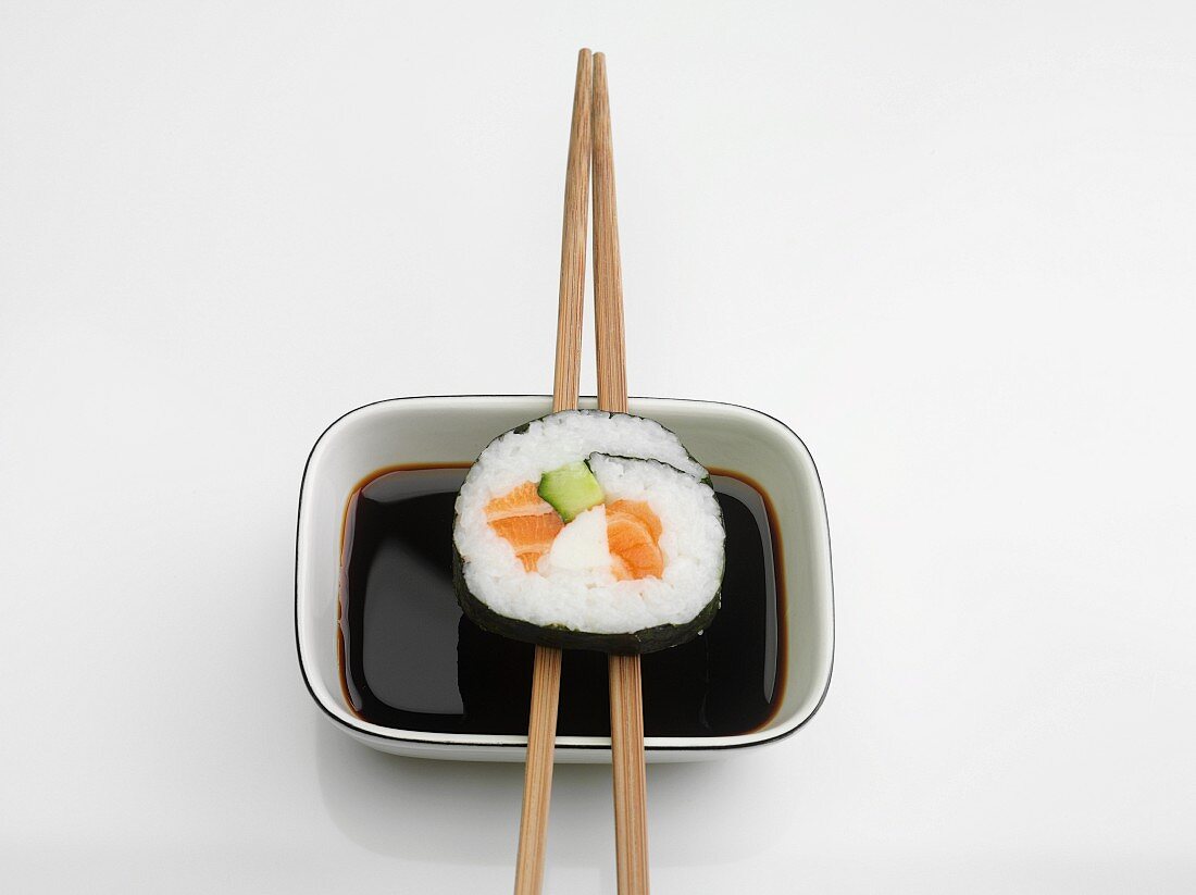 A maki sushi roll with salmon and cucumber balanced on chopsticks over a dish of soy sauce