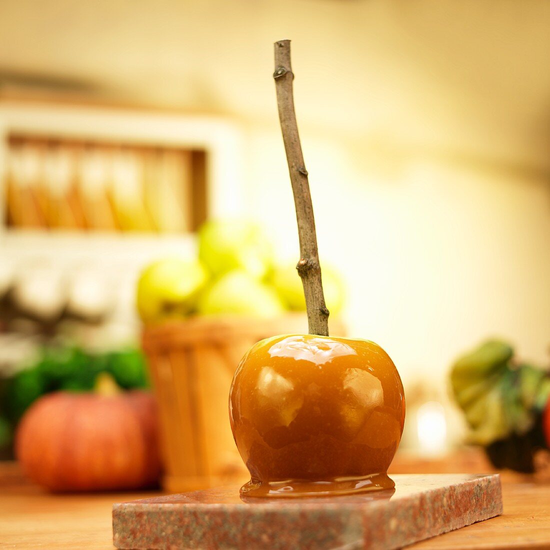 A caramel apple with a stick in front of apples and pumpkins in a kitchen