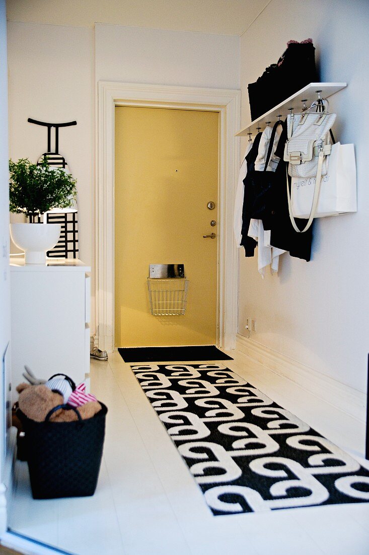 Schwarz-weiss gemusterter Läufer auf weißem Boden im Eingangsbereich, Wandgarderobe, neben hellgelber Wohnungstür mit Korb für Briefe