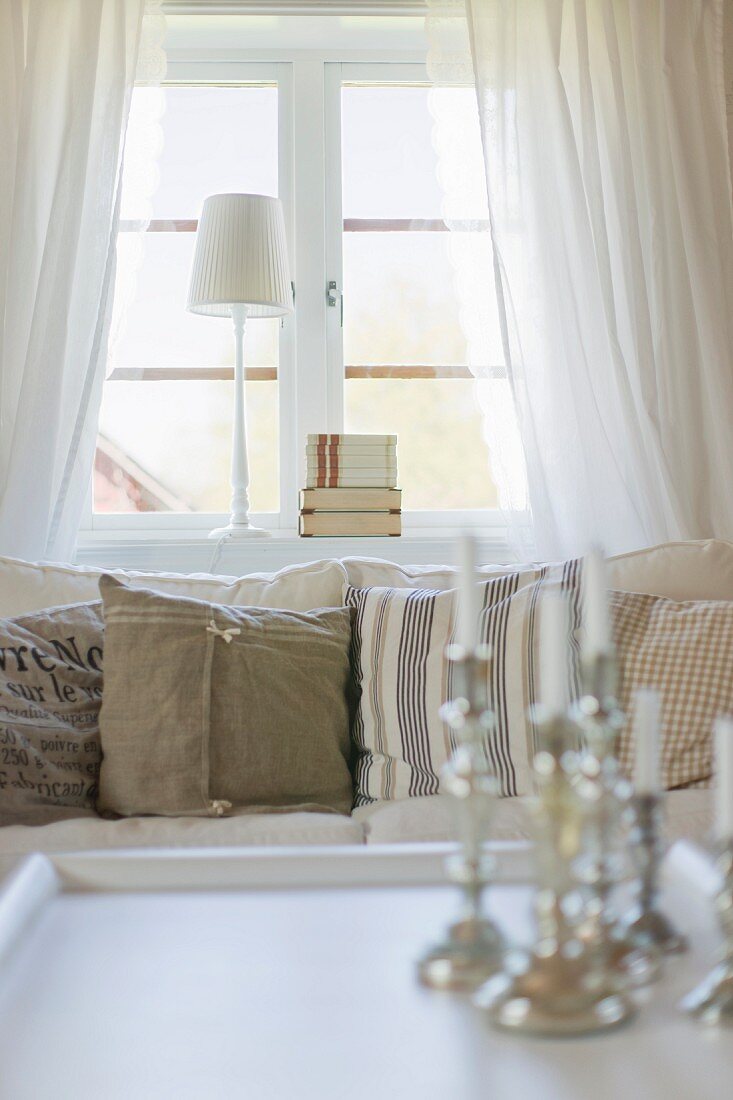 View across table to row of country-house-style scatter cushions on sofa and table lamp on windowsill