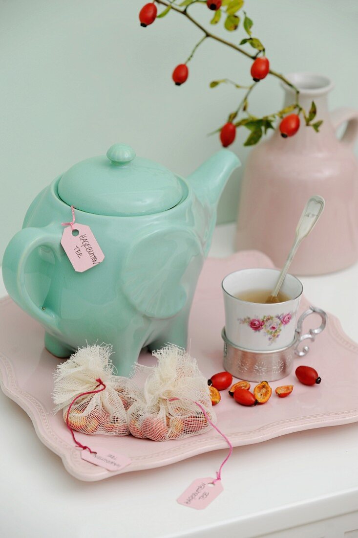 Türkisgrüne Teekanne und Vintage Tasse mit Hagebuttenhälften und Teesäckchen auf rosafarbenem Serviertablett