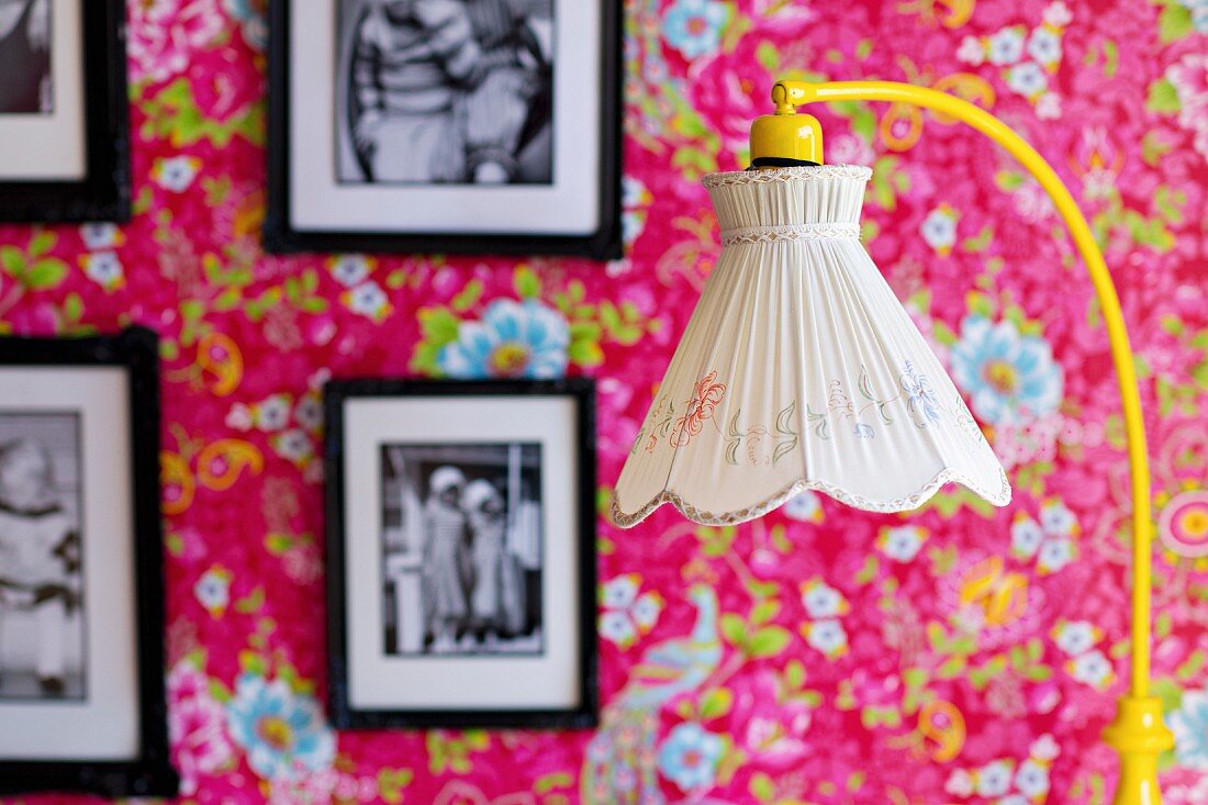 Standard lamp with nostalgic lampshade in front of framed black and white photos on floral wallpaper