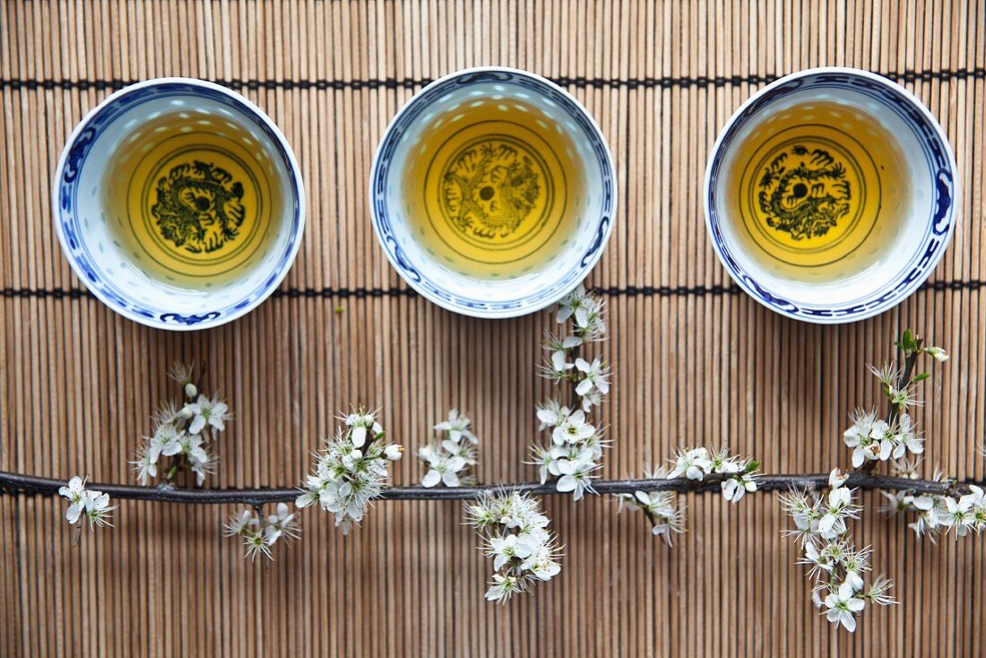 Drei asiatische Teeschälchen mit Grüntee und Kirschblütenzweig auf Bambusmatte