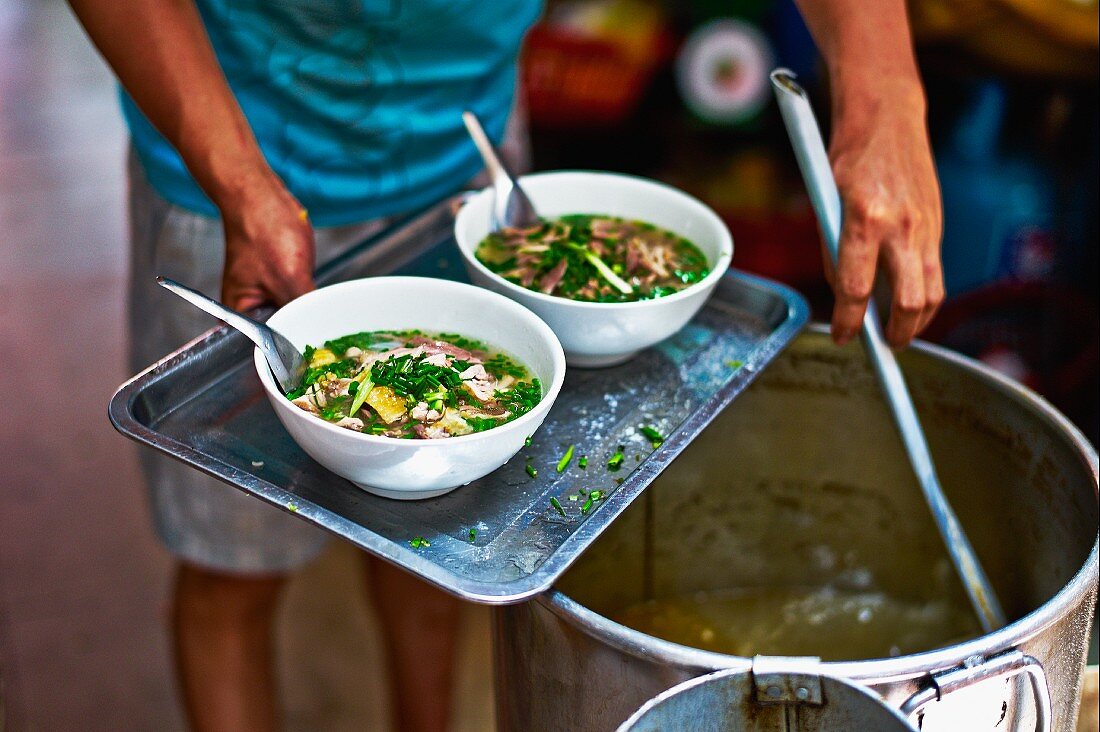 A man ladling pho ga (Vietnames chicken soup) from a vat into bowls