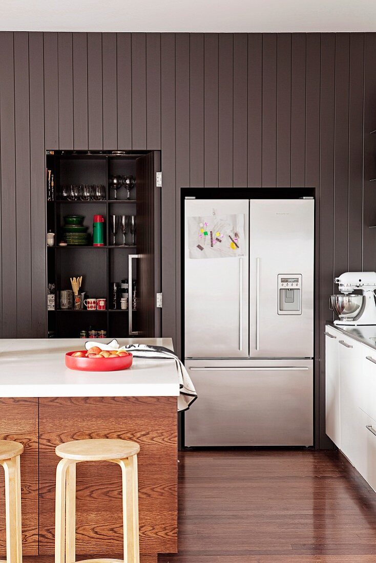 Island counter with wooden stools in front of black cupboard with folding door and fridge built into grey wall panelling