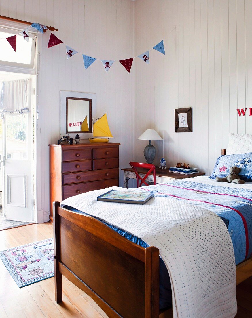 Bed with patchwork bedspread, bunting above wooden chest of drawers and white wood-panelling in cheerful, country-house, child's bedroom