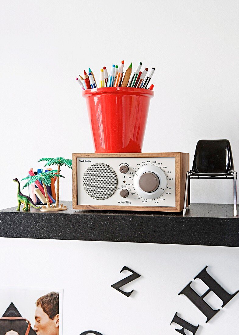 Coloured pencils in red pot in top of radio flanked by miniature chair and toys on black, wall-mounted shelf