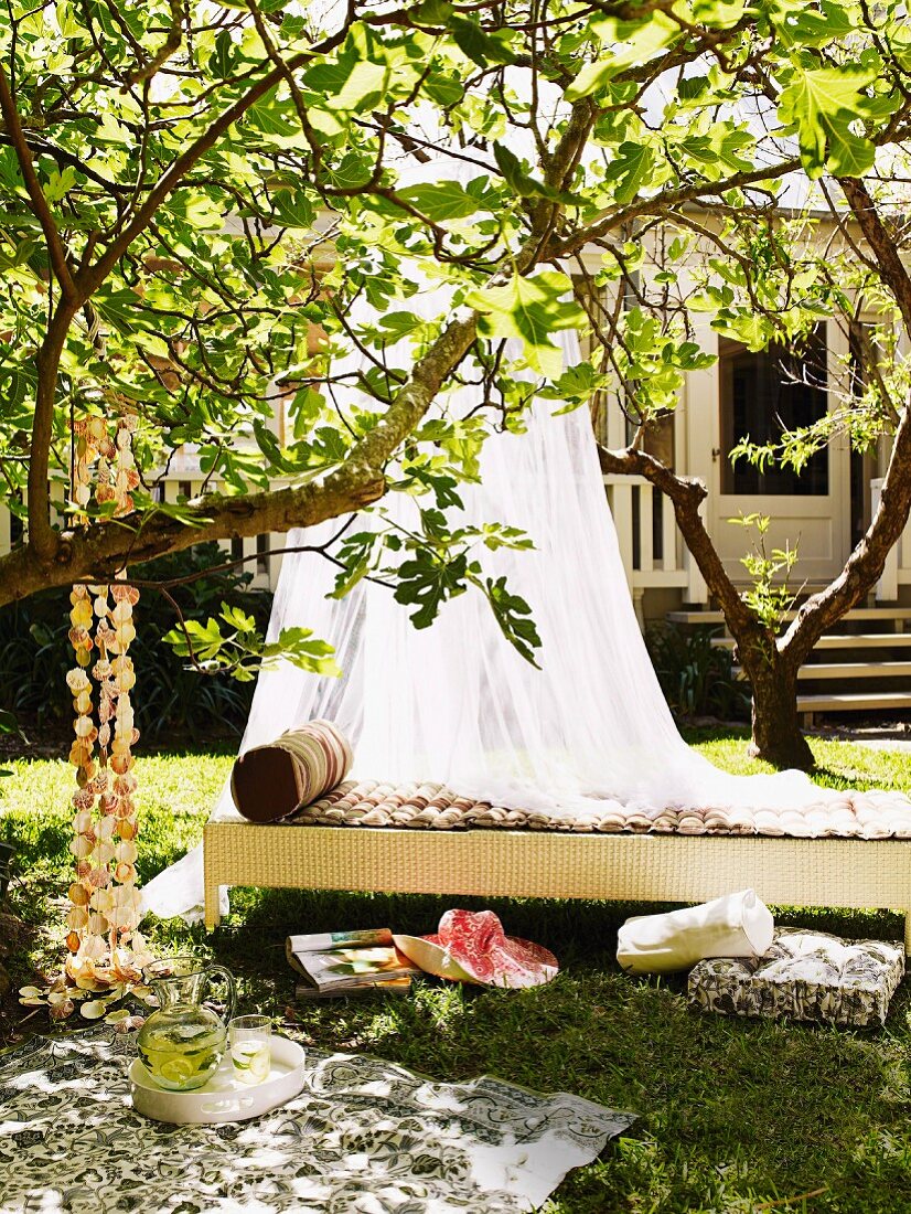 Shady spot in garden - airy canopy above wicker day bed behind picnic blanket and cushions on lawn