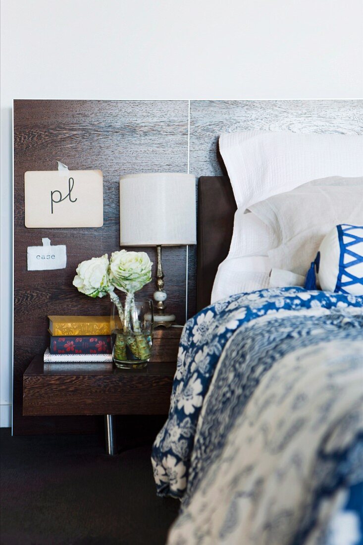 Bed with blue and white bed linen, bedside cabinet and dark wood headboard; calligraphy on paper taped to headboard, vase of flowers and antique lamp