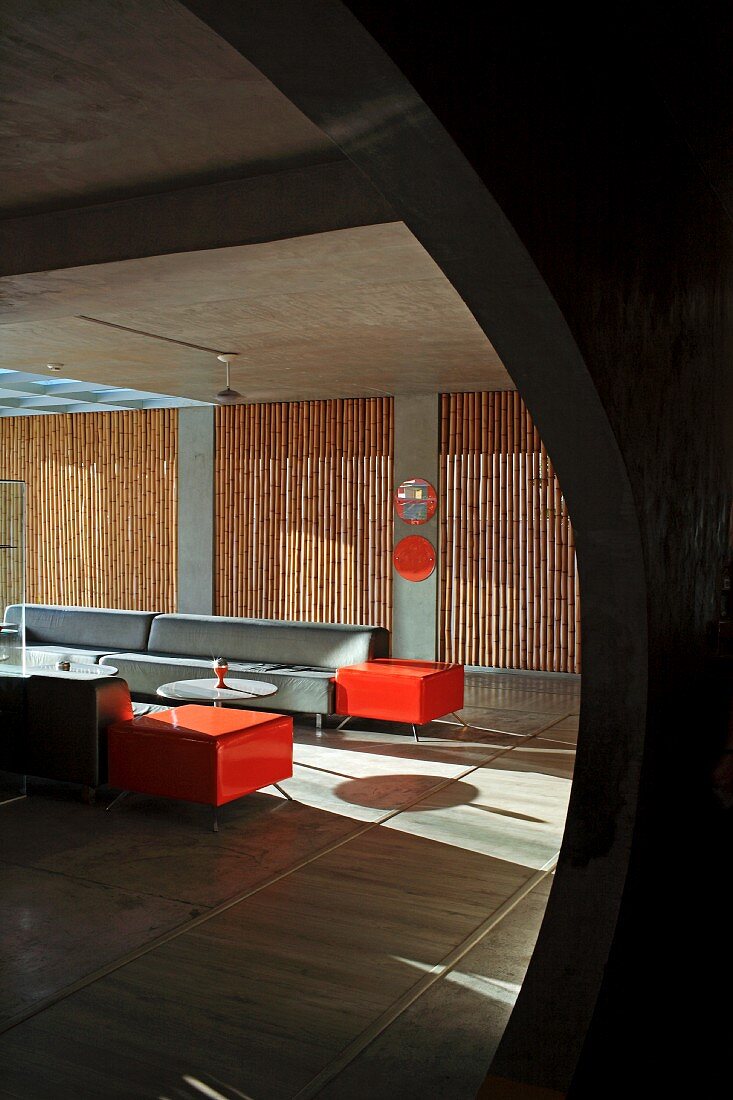 Blick durch kreisförmige Öffnung in minimalistischem Lounge-Bereich mit Sofagarnitur in Grau und Orange, im Hintergrund Bambusstäbe in Fensternischen als Sichtschutz