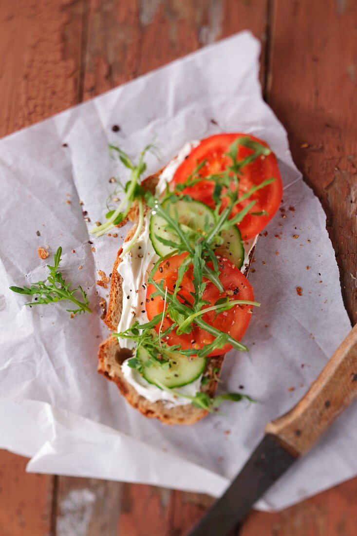 A slice of bread topped with cream cheese, cucumber, tomato and rocket
