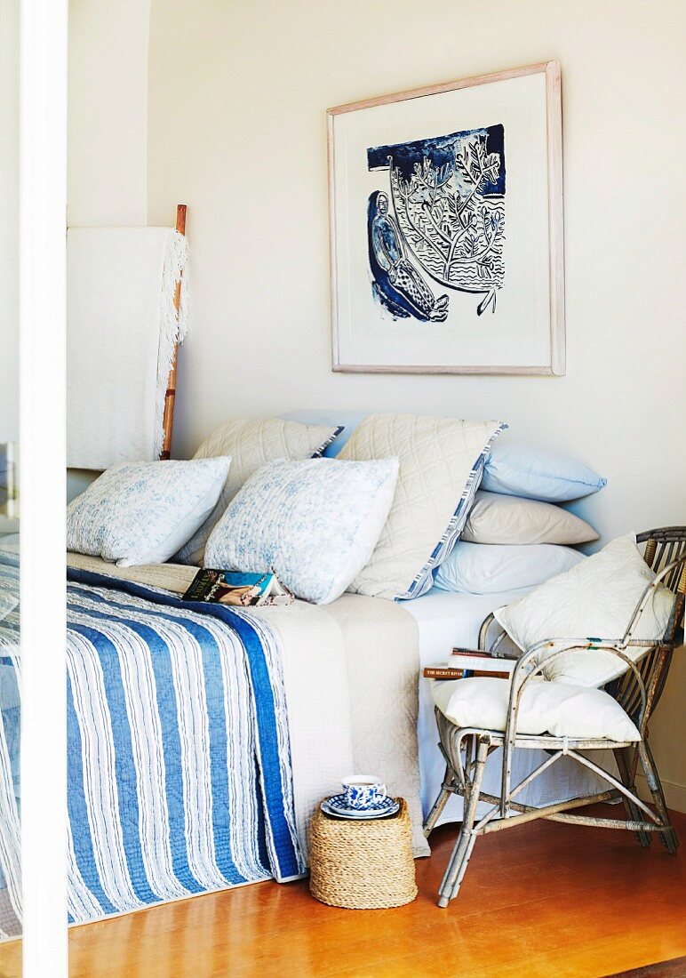 Wicker chair next to French bed with stacks of pillows and blue striped bedspread below framed picture on wall