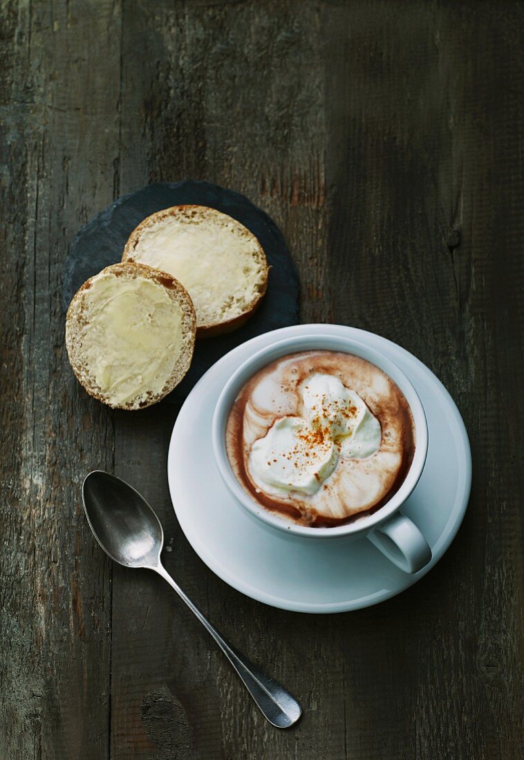 A buttered roll and a cup of hot chocolate with milk foam and cream