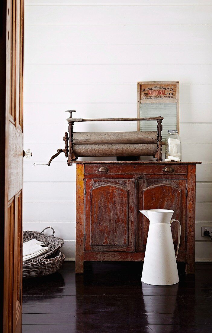 White metal water jug on dark wooden floor in front of old mangle on top of antique wooden cabinet