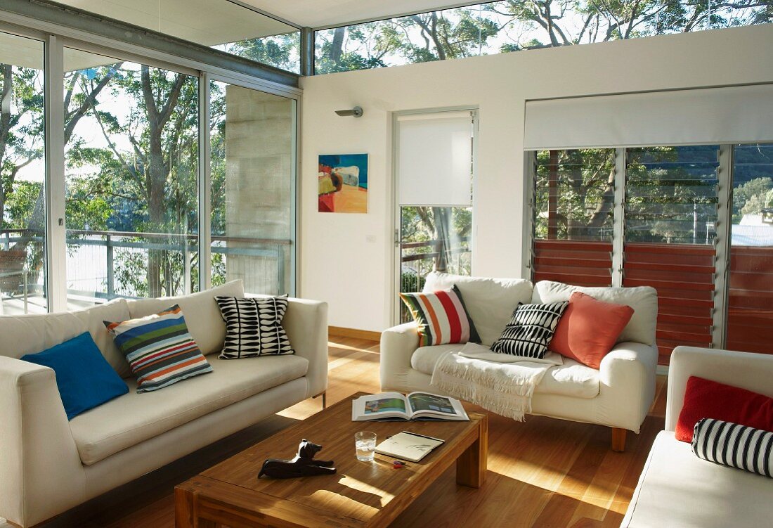 White sofa set with scatter cushions and simple wooden coffee table in interior with glass walls