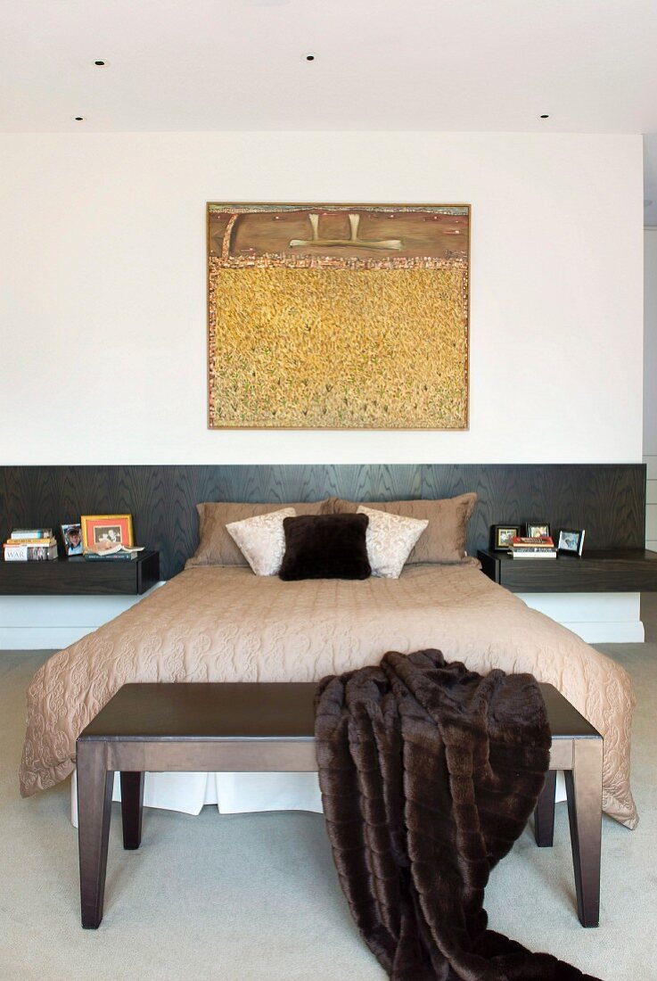 Moderne Kunst über französischem Bett mit schwarzem Kopfteil und integrierten Nachtischen; Felldecke auf Kleiderbank im Vordergrund