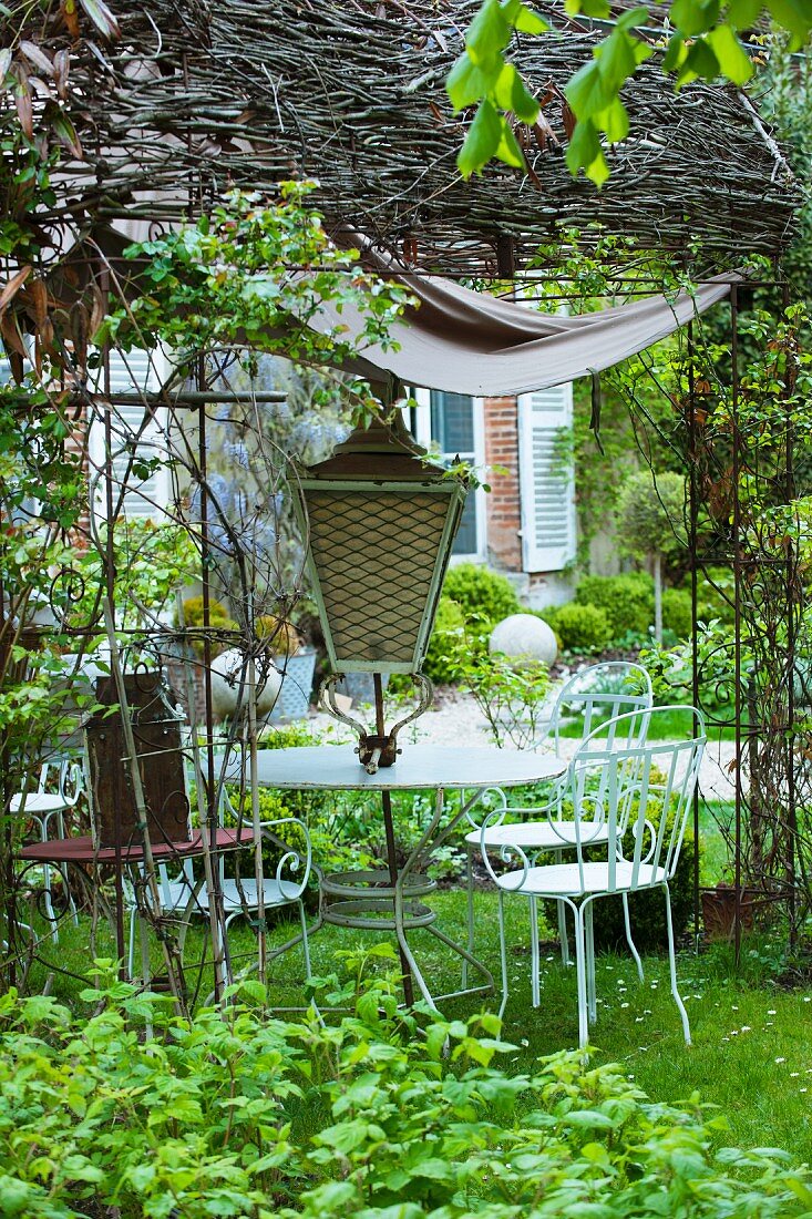 A white-painted metal table and garden chairs for sale with an antique lantern under a wicker pergola