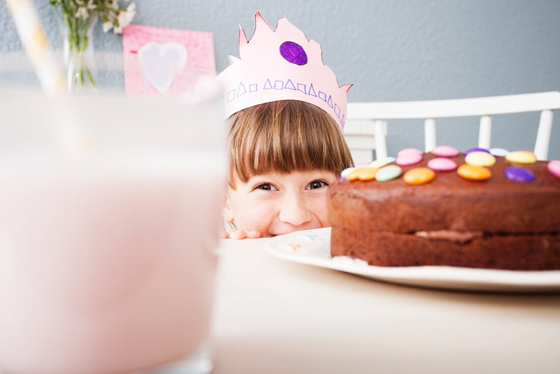A little girl wearing a crown hiding behind a decorated chocolate cake