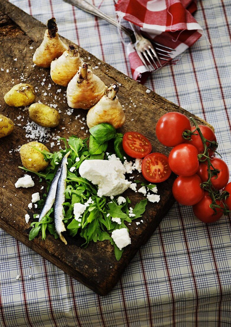 Holzbrett mit gebratenen Hühnerkeulen, Kartoffeln, Rucola, Tomaten und Schafskäse