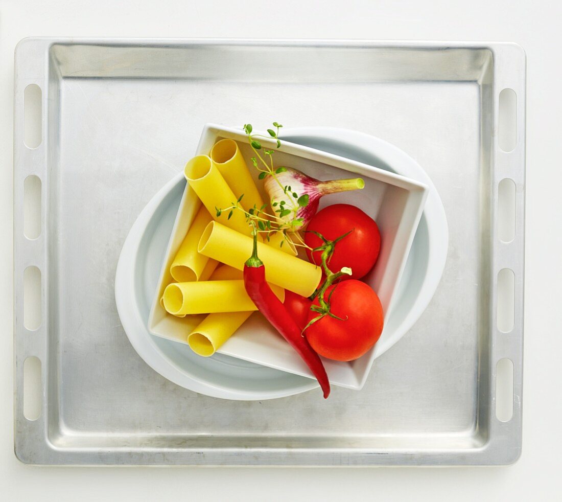 Pasta, a chilli pepper, tomatoes and garlic in a baking dish on a baking tray