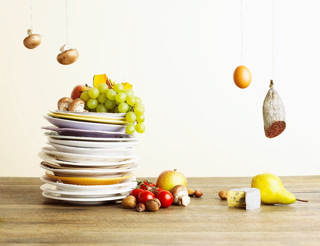 A stack of plates with fruit, vegetables and mushrooms next to an egg, salami and cheese