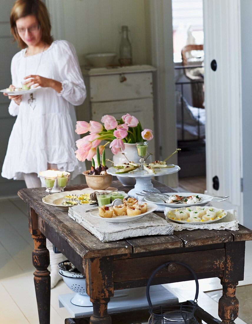 A festive buffet with various canapés and a bunch of tulips on a rustic wooden table with a young woman eating in the background