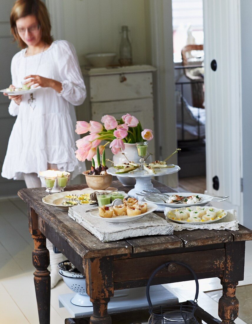 Festliches Buffet mit verschiedenen Häppchen und Tulpenstrauss auf rustikalem Holztisch; im Hintergrund eine essende junge Frau