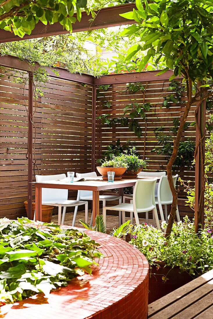 Kreisförmiges Hochbeet mit roten Mosaikfliesen, im Hintergrund lauschiger Sitzplatz vor elegantem Sichtschutzzaun einer Terrasse