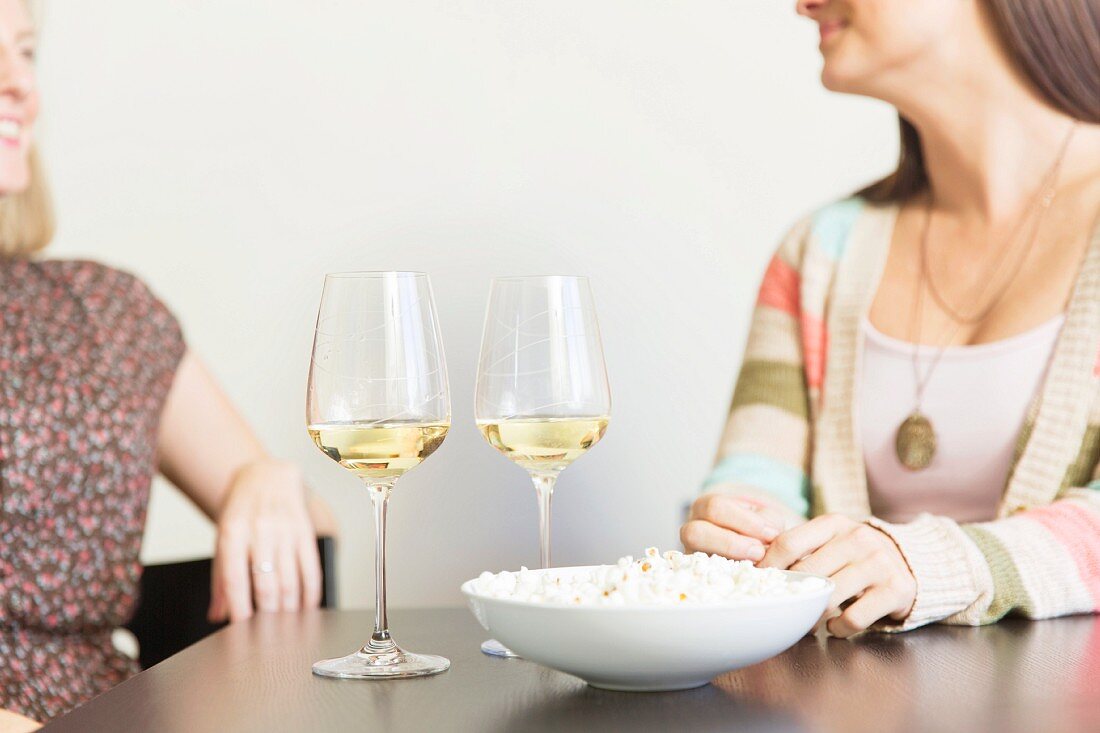 Two women sitting at a table with a bowl of popcorn and glasses of wine