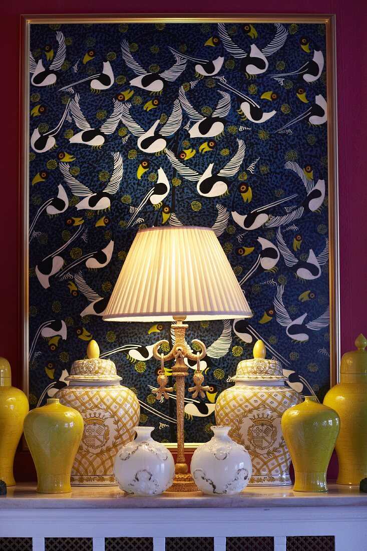 Tischleuchte mit hellem Plissee Lampenschirm, Sammlung aus Keramikvasen in Gelb und Behälter mit Deckel in Gelb Weiß vor Bild mit Vogelmotiven