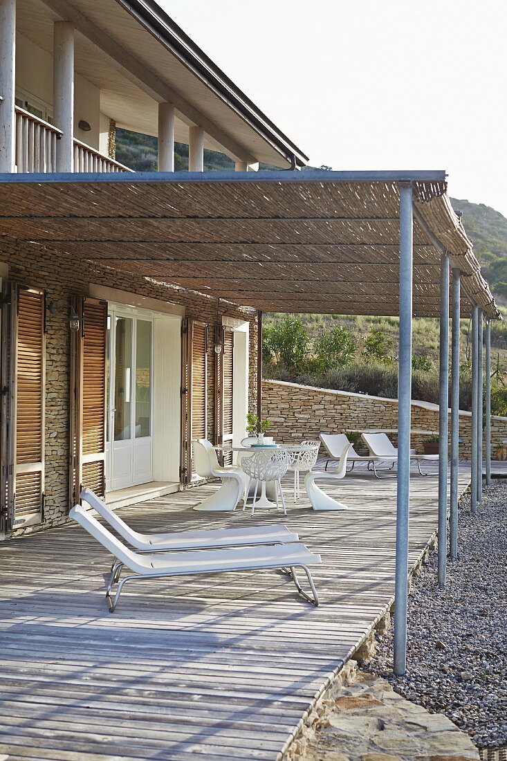 Sun loungers and white outdoor furniture on wooden deck with bamboo pergola adjoining Mediterranean house