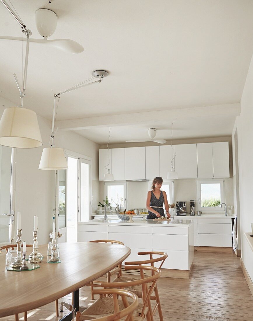 Dining area with classic wooden chairs, adjustable pendant lamps with white lampshades; woman in open-plan, modern kitchen in background