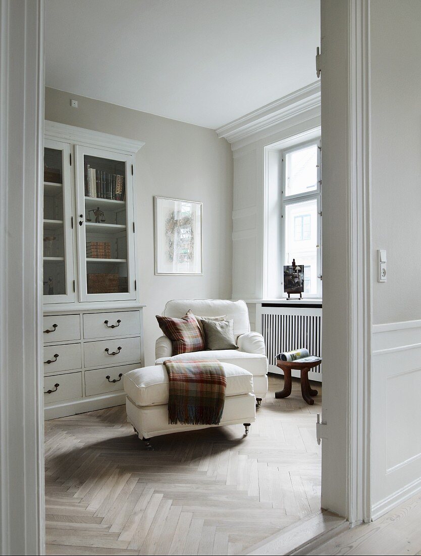 View through open door of white armchair with matching footstool next to dresser with glass-fronted top in traditional interior