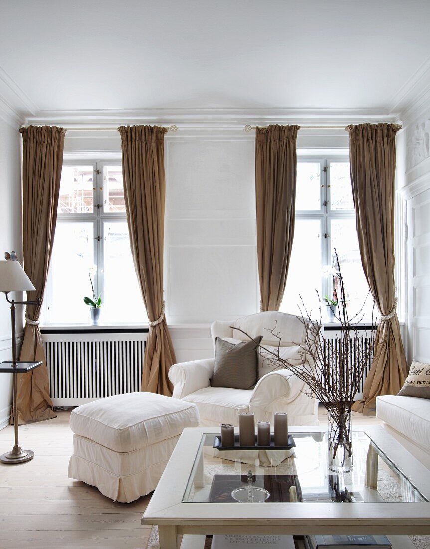 Vase of branches on glass-topped coffee table in front of white armchair with matching footstool and windows with draped, light brown curtains