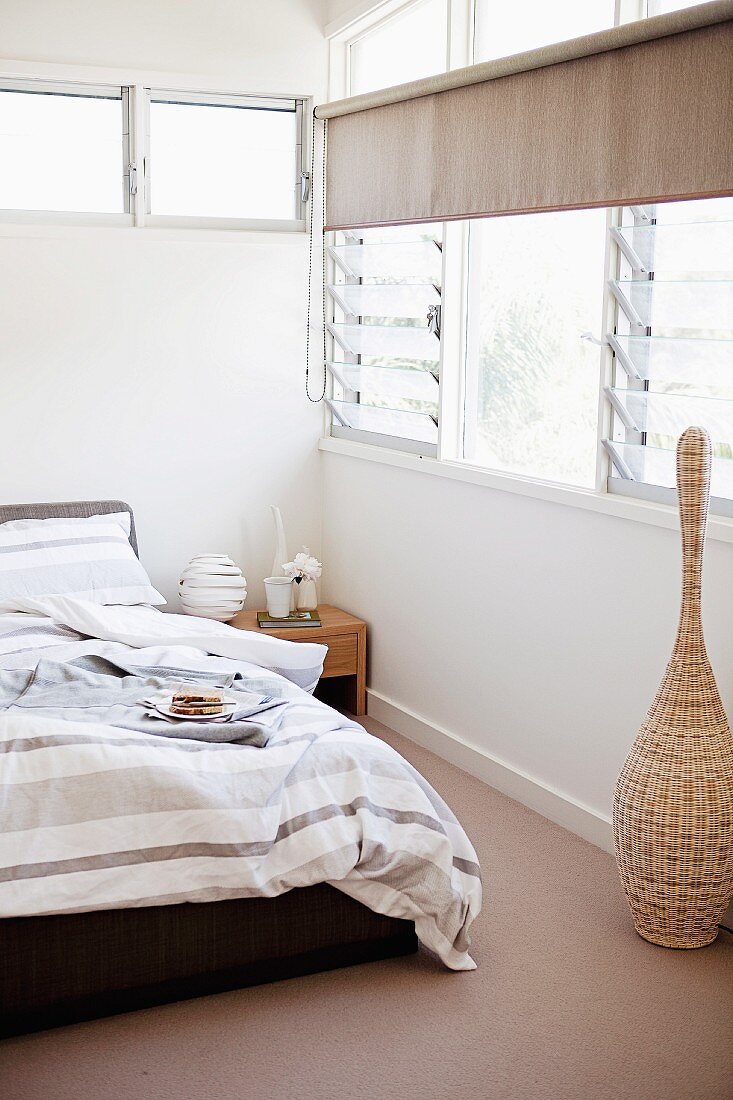 Bed with striped bed linen in shades of grey and brown in corner of bedroom with louvre windows and transom window