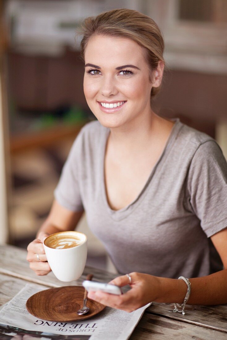 Junge Frau mit Cappuccino, Handy und Tageszeitung