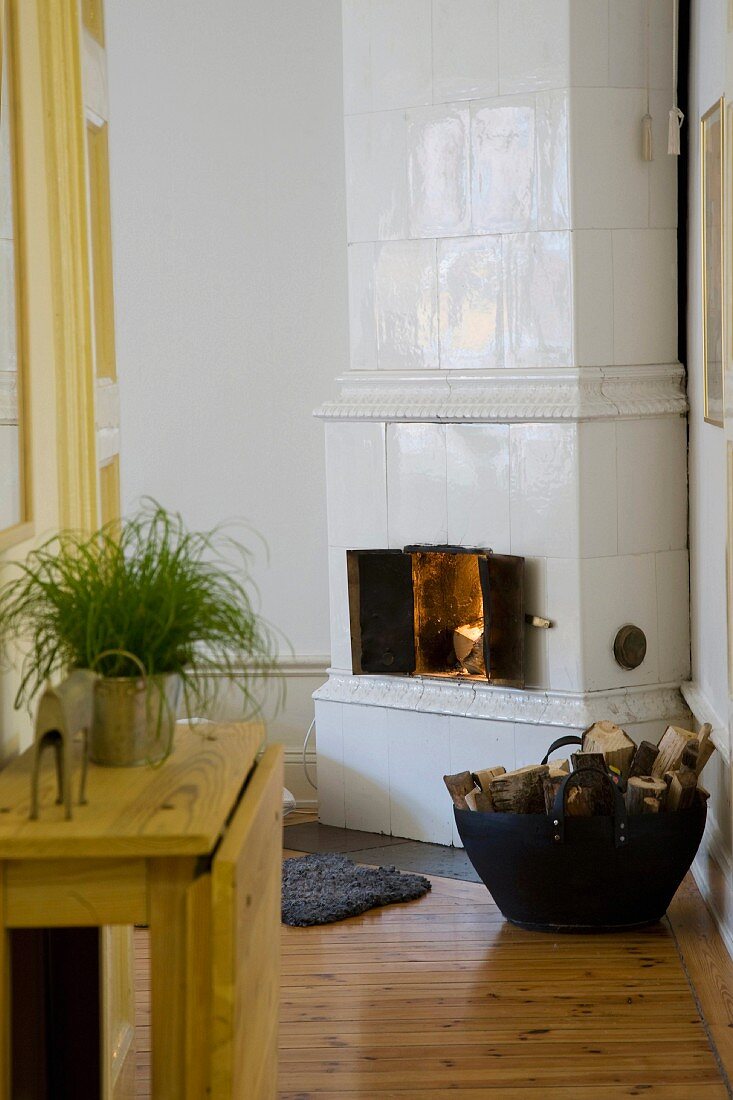 Tiled stove clad in white tiles and basket of firewood on board floor