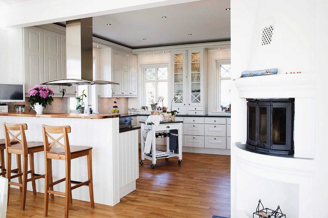 Wooden bar stools at breakfast bar integrated into kitchen counter below stainless steel extractor hood in open-plan, white country-house kitchen