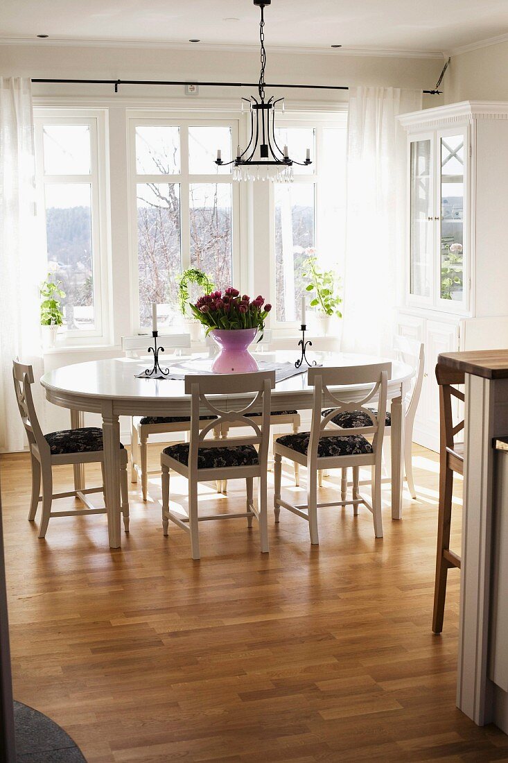 Oval dining table and white wooden chairs below wrought iron chandelier in front of window