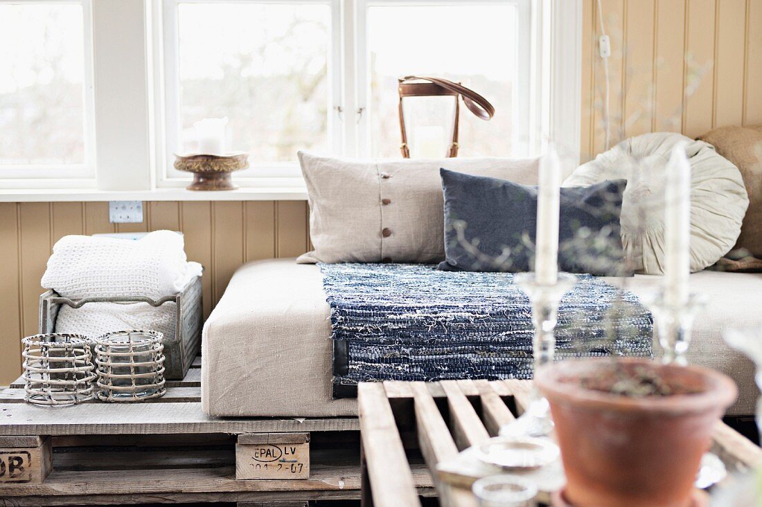 View across coffee table to couch made from wooden pallets and pale cushions below window