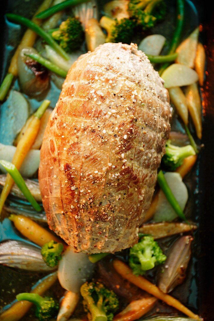 A whole roast pork roulade on a bed of vegetables in a roasting tin