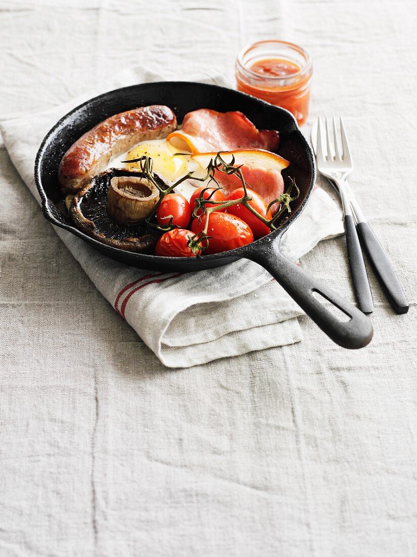 And English breakfast featuring bacon, sausage, fried egg, Portobello mushrooms and tomatoes in a pan