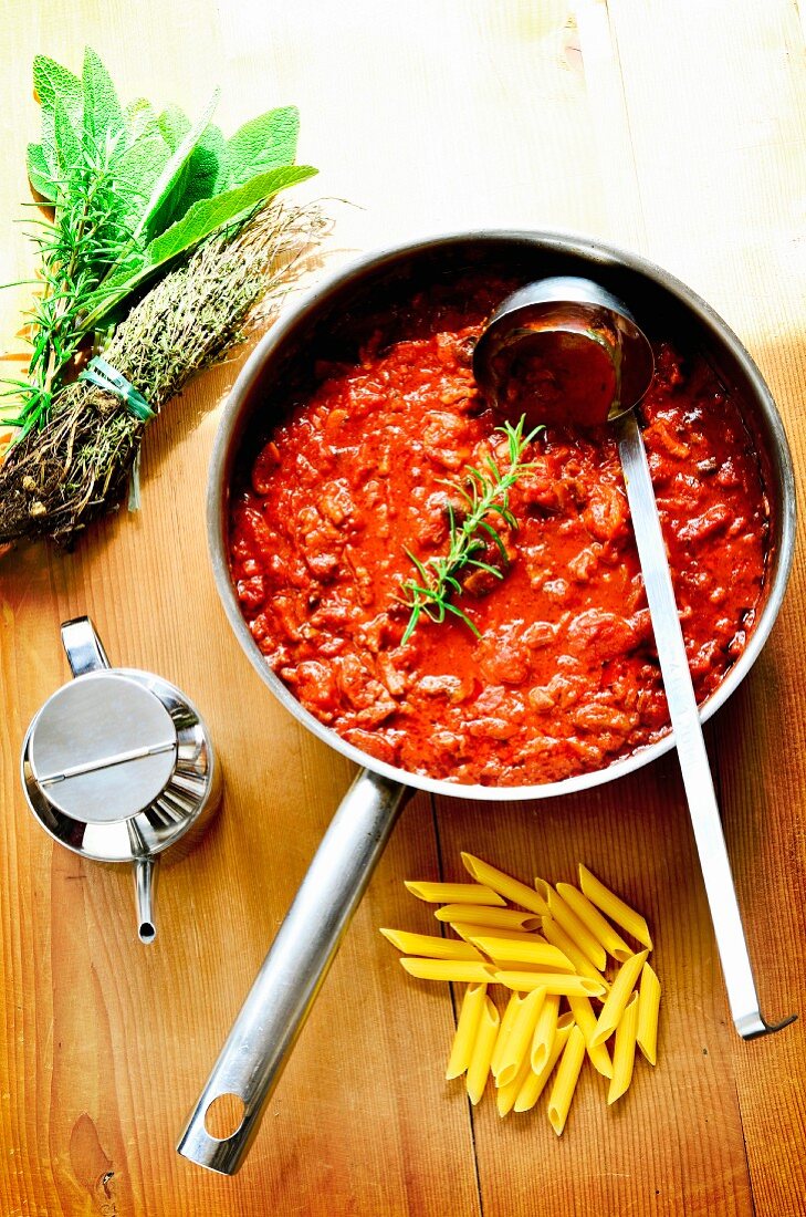 Sauce Bolognese in Pfanne auf Holzbrett, Penne, Kräuter und Olivenöl