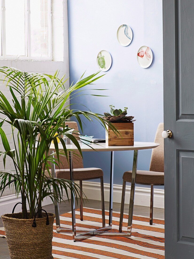 View through open door of potted palm in dining area and upholstered chairs around table with chrome frame in corner