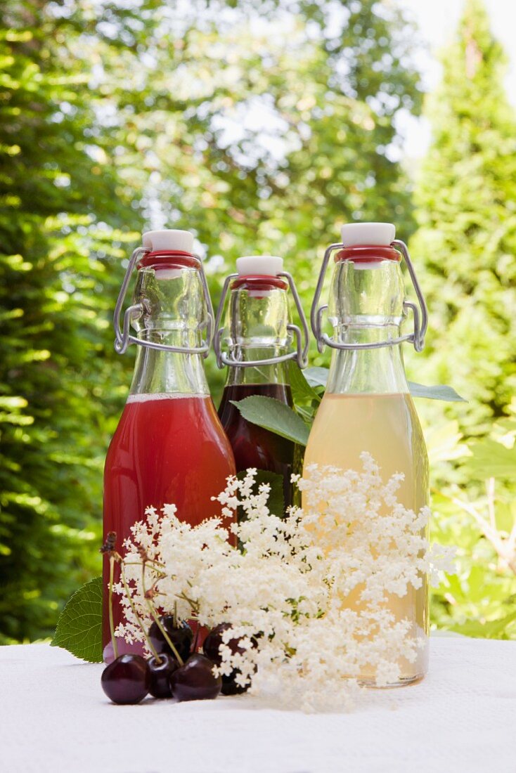 Drei Glasflaschen selbstgemachter Sirup auf Gartentisch: Rhabarbersirup, Kirschsirup, Holunderblütensirup