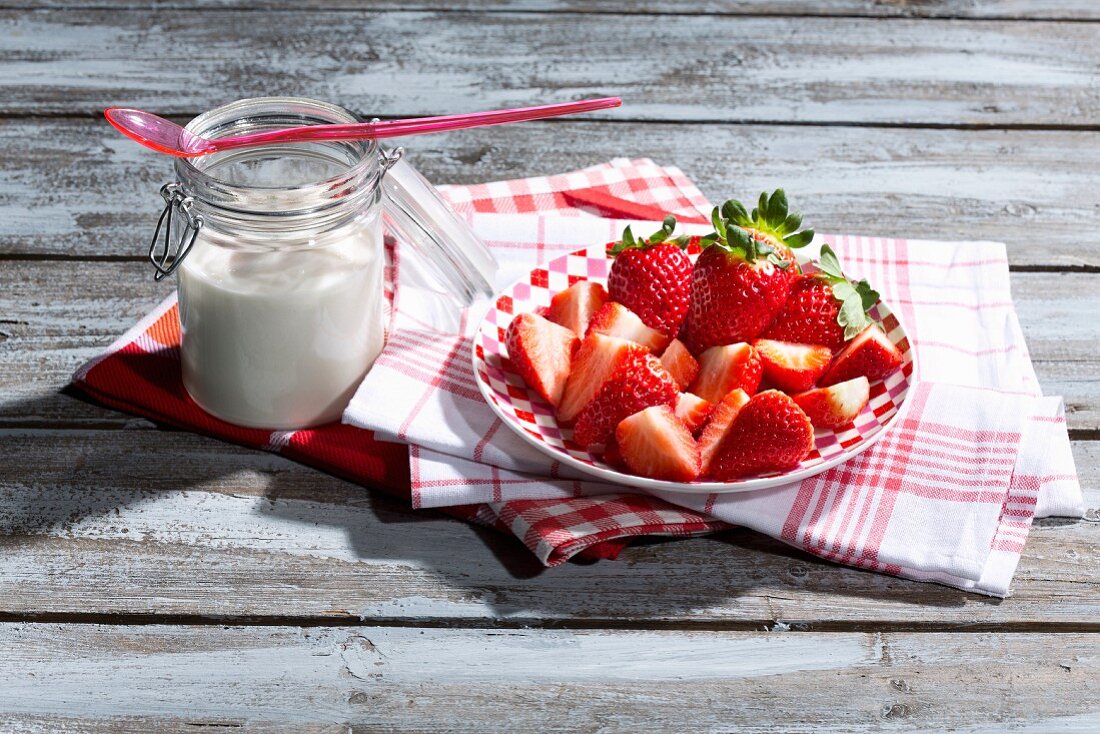 Ein Glas Naturjoghurt und Teller mit Erdbeeren auf Küchentüchern und Holztisch
