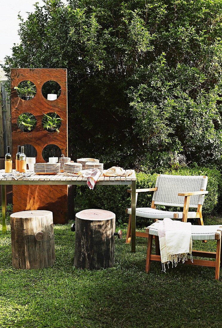 Set table, tree stump stools and armchair in garden; wooden panel with potted plants in circular apertures in background