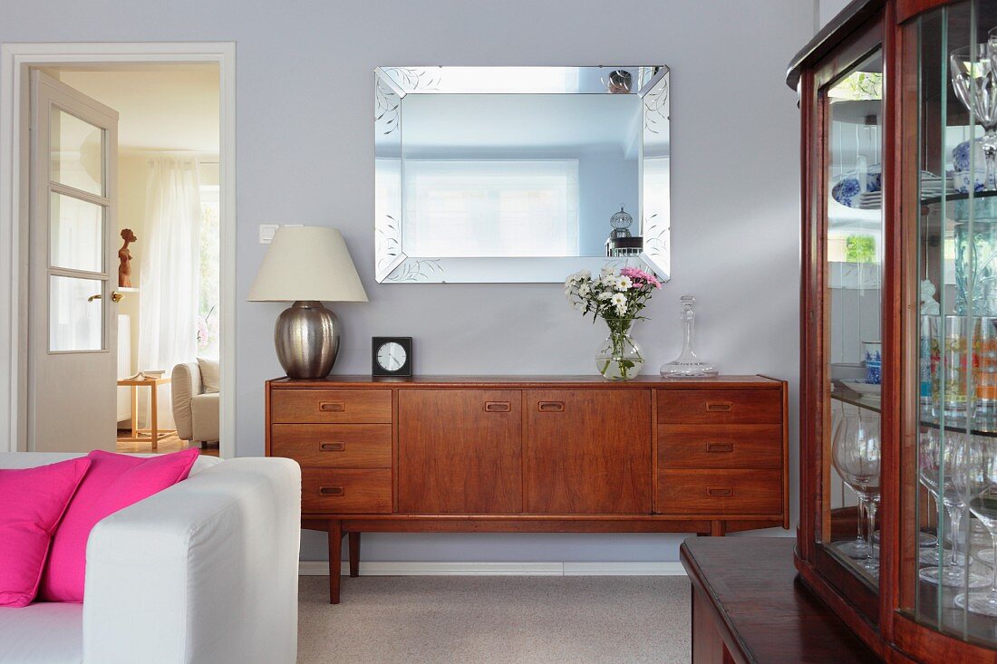 Fifties-style wooden sideboard against wall, white sofa with pink scatter cushions and antique, glass-fronted cabinet